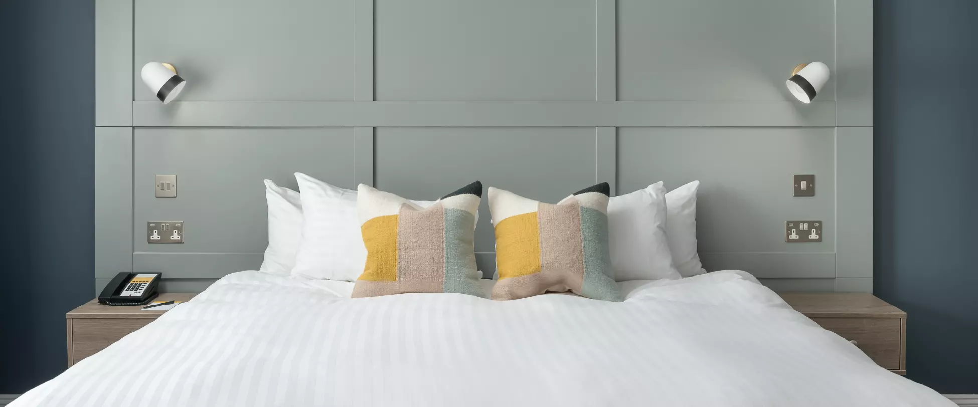 Standing at the foot of a large bed the photograph shows the head end of the bed with lamps and tables on either side. There are cushions and pillows at the head of the bed..